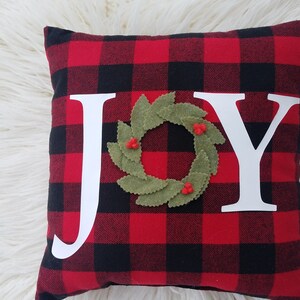 May include: Red and black buffalo plaid pillow with the word "JOY" in white letters. A green felt wreath with red berries is centered between the "O" and the "Y".
