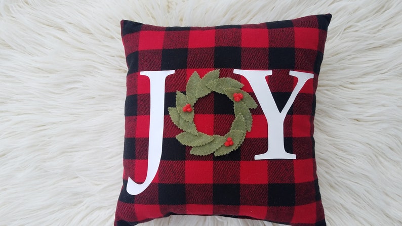 May include: Red and black buffalo plaid pillow with the word "JOY" in white letters. A green felt wreath with red berries is centered between the "O" and the "Y".