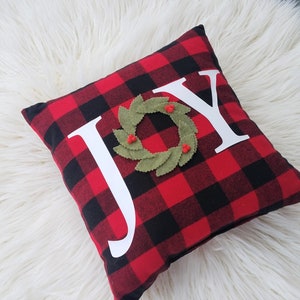 May include: Red and black buffalo plaid pillow with the word "JOY" in white letters. A green felt wreath with red berries is centered on the pillow.