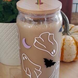 May include: A glass jar with a wooden lid and a straw sticking out of the top. The jar is filled with a light brown liquid and has a Halloween-themed design with ghosts and bats.