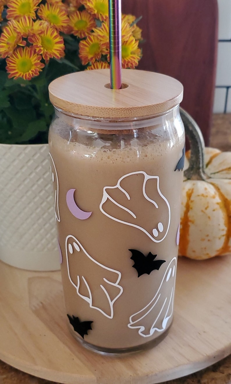 May include: A glass jar with a wooden lid and a straw sticking out of the top. The jar is filled with a light brown liquid and has a Halloween-themed design with ghosts and bats.
