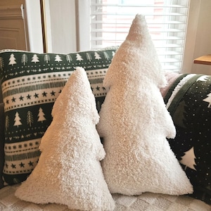 Puede incluir: Dos almohadas de árbol de Navidad de felpa blanca en un sofá beige con una almohada estampada verde y blanca detrás.