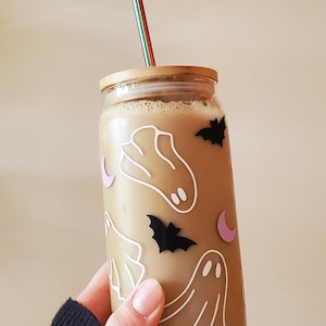 May include: A glass jar with a bamboo lid and a rainbow-colored metal straw. The jar is filled with a light brown liquid and decorated with white ghost and bat stickers.