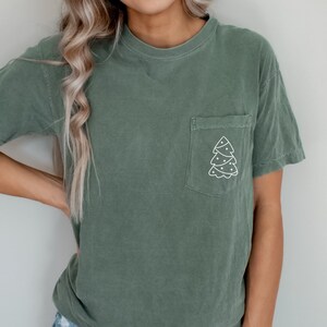 May include: Sage green t-shirt with a pocket featuring a white Christmas tree design. The short-sleeved shirt is made of a soft material and has a relaxed fit. A casual top for the holiday season.
