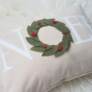 May include: A beige decorative pillow with the word "NOEL" in white block letters. A green felt wreath with red berry accents is centered on the pillow, creating a festive holiday theme.