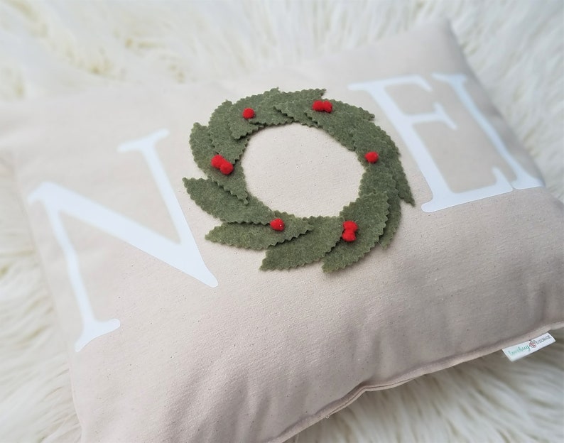 May include: A beige decorative pillow with the word "NOEL" in white block letters. A green felt wreath with red berry accents is centered on the pillow, creating a festive holiday theme.