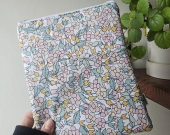 Op de afbeelding: Een tablet hoes met een bloemenpatroon op een witte achtergrond, met roze, gele en blauwe bloemen en groene bladeren. De hoes wordt vastgehouden door een persoon met een zwarte mouw. De hoes heeft een ritssluiting.