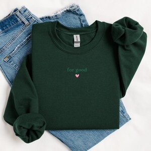 May include: A dark green sweatshirt with the text "for good" and a small pink heart embroidered on the front.