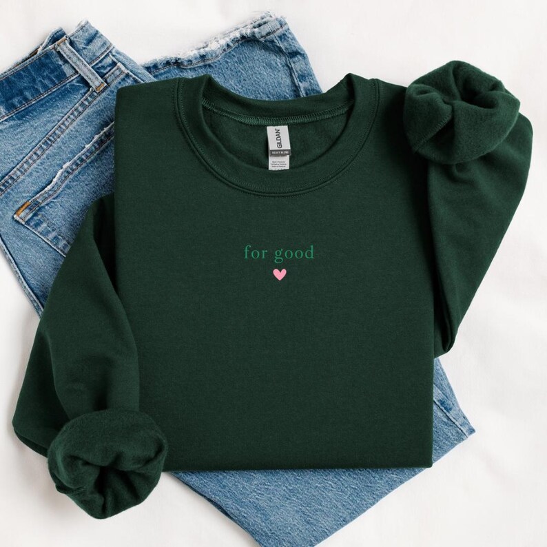 May include: A dark green sweatshirt with the text "for good" and a small pink heart embroidered on the front.