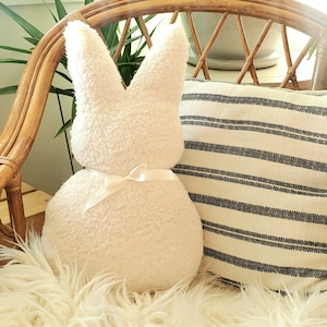 May include: A white plush bunny rabbit pillow with a white satin bow. The pillow is sitting on a white fluffy rug in a wicker chair.
