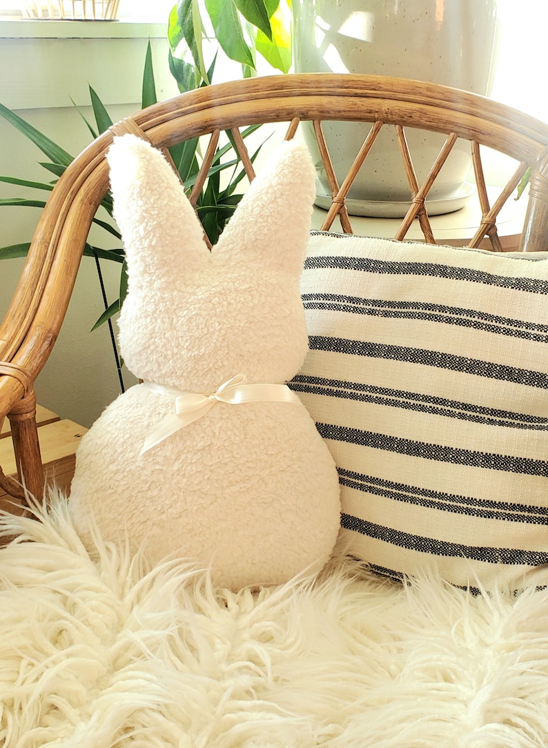 May include: A white plush bunny rabbit pillow with a white satin bow. The pillow is sitting on a white fluffy rug in a wicker chair.