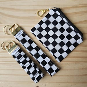 May include: A collection of black and white chequered accessories, including a small zipped pouch, two keychains, and a wristlet. The keychains and wristlet have gold-coloured hardware. The items are displayed on a light brown wooden surface.