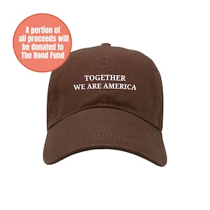 Puede incluir: Gorra de béisbol marrón con el texto blanco "TOGETHER WE ARE AMERICA". Una parte de los beneficios se donará a The Bond Fund. La gorra tiene una visera curva y un cierre de correa de tela.