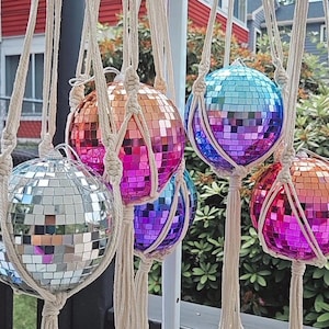 May include: Five disco ball ornaments hanging from macrame string. The disco balls are silver, pink, blue, and purple. The macrame is white.