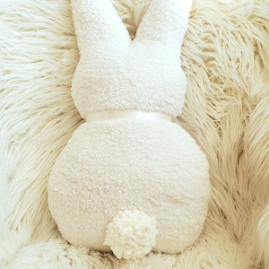 May include: A white plush bunny rabbit pillow with a white pom-pom tail. The pillow is sitting on a white fluffy surface.