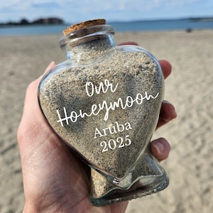 May include: A heart-shaped glass bottle filled with sand, sealed with a cork. The bottle has the words "Our Honeymoon Aruba 2025" in white script. The background is a beach with the ocean in the distance.