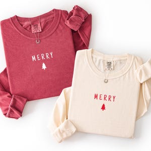 Puede incluir: Dos camisetas de manga larga, una burdeos y otra crema, con la palabra "MERRY" y un gráfico de árbol de Navidad. Cada camiseta tiene cuello redondo y un collar plateado. La camiseta burdeos tiene un gráfico blanco, mientras que la crema tiene un gráfico rojo.