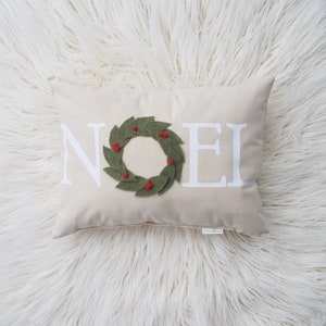 May include: A rectangular beige pillow with the word "NOEL" in white block letters. A green felt wreath with red berry accents is centered on the pillow. The pillow is on a white, fluffy surface.