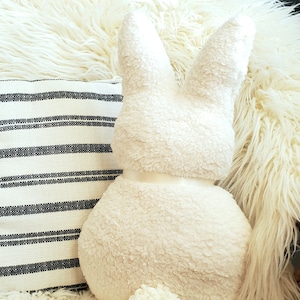 May include: A white plush bunny rabbit pillow with a fluffy tail. The pillow is sitting on a white fluffy rug.