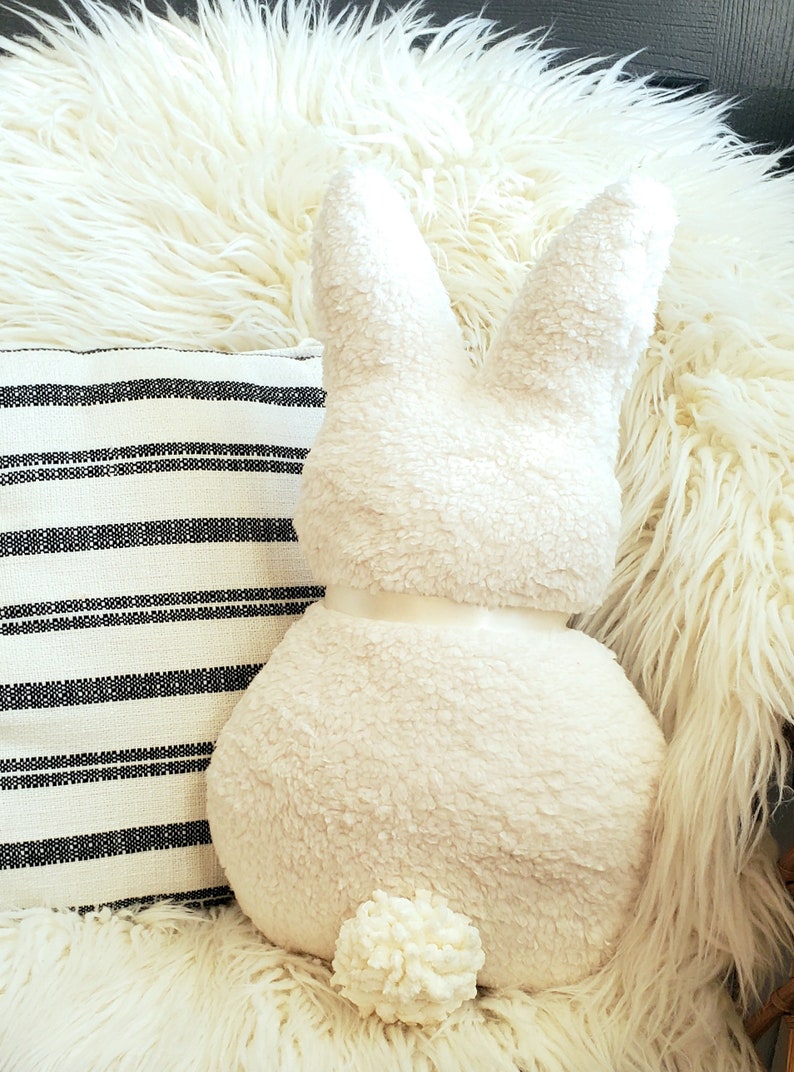 May include: A white plush bunny rabbit pillow with a fluffy tail. The pillow is sitting on a white fluffy rug.