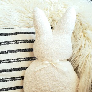 May include: A white plush bunny pillow with a white satin bow. The pillow is sitting on a white fluffy rug.