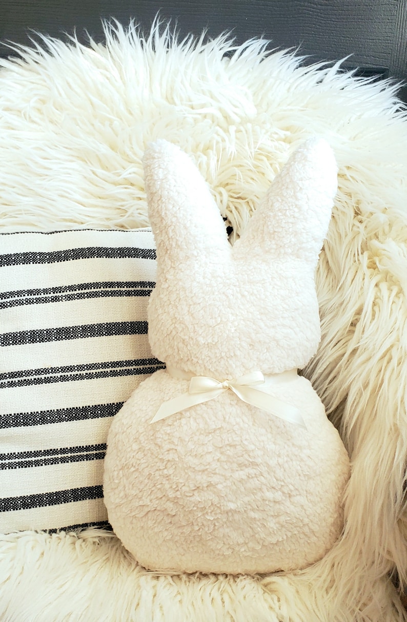 May include: A white plush bunny pillow with a white satin bow. The pillow is sitting on a white fluffy rug.