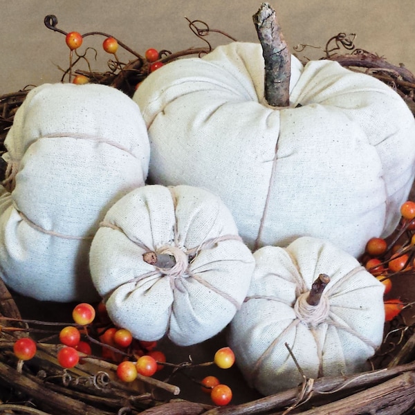 May include: Four off-white fabric pumpkins of varying sizes are nestled within a dark brown woven grapevine wreath. Each pumpkin features a natural twig stem and is wrapped with thin twine. Small orange berries are scattered throughout.