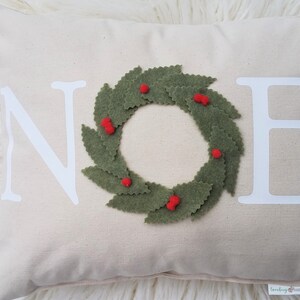 May include: A rectangular beige pillow with the word "NOEL" in white block letters. A green felt wreath with red berry accents is centered on the pillow. The pillow is on a white, fluffy surface.