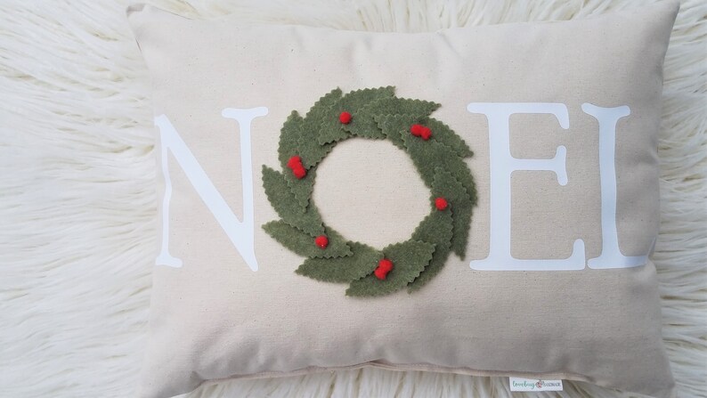 May include: A rectangular beige pillow with the word "NOEL" in white block letters. A green felt wreath with red berry accents is centered on the pillow. The pillow is on a white, fluffy surface.