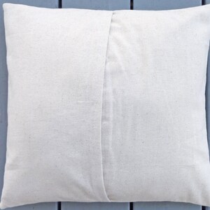 May include: A square, off-white pillow cover made of a textured fabric. The back of the pillow features a seam down the center. The pillow is likely for home decor.