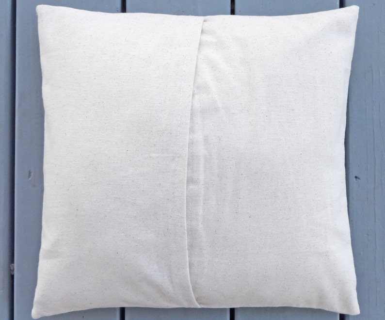 May include: A square, off-white pillow cover made of a textured fabric. The back of the pillow features a seam down the center. The pillow is likely for home decor.