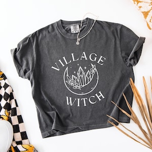 May include: A charcoal gray short-sleeve t-shirt featuring a white graphic print of a crescent moon, crystals, and flowers with the text "VILLAGE WITCH" arched around the design. A thin silver-toned chain necklace rests on the neckline.