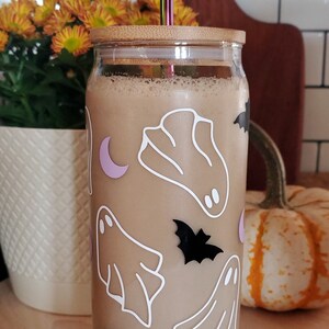 May include: A glass reusable tumbler with a bamboo lid and a straw. The tumbler is filled with a brown liquid and has a Halloween theme with white ghosts and black bats.