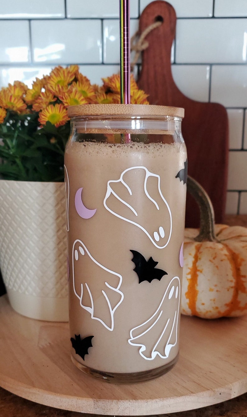 May include: A glass reusable tumbler with a bamboo lid and a straw. The tumbler is filled with a brown liquid and has a Halloween theme with white ghosts and black bats.