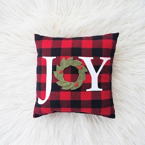 May include: Red and black plaid pillow with the word "JOY" in white letters. The "O" is a green felt wreath with red felt berries.