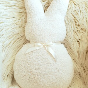 May include: A white plush bunny pillow with a white satin ribbon tied around the middle. The pillow is sitting on a white fluffy surface.