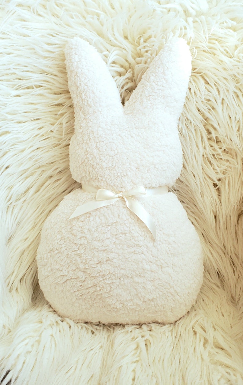 May include: A white plush bunny pillow with a white satin ribbon tied around the middle. The pillow is sitting on a white fluffy surface.