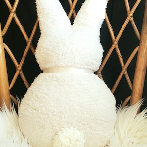May include: A white plush bunny rabbit pillow with a white satin bow and a fluffy white tail. The pillow is sitting on a white fluffy chair.