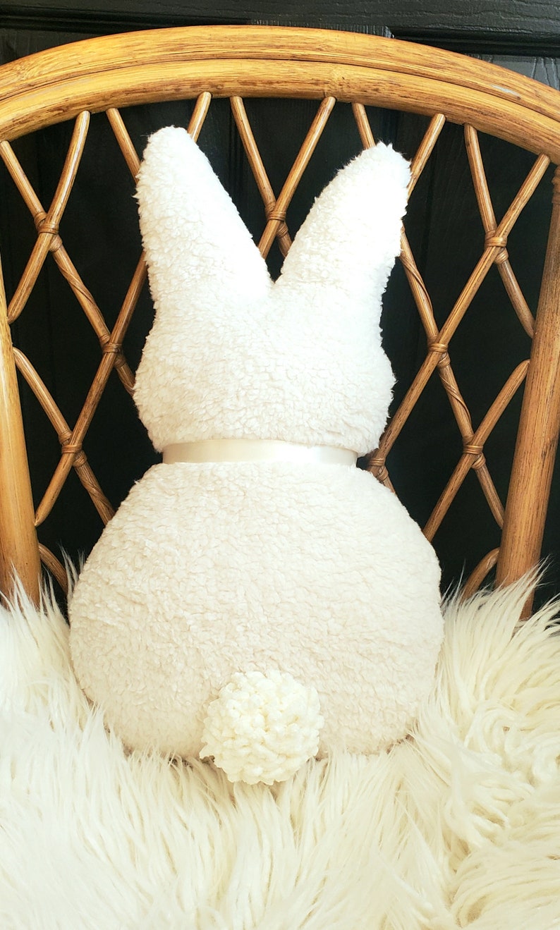May include: A white plush bunny rabbit pillow with a white satin bow and a fluffy white tail. The pillow is sitting on a white fluffy chair.