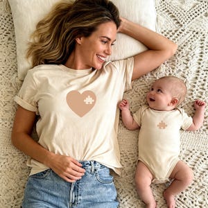 May include: A beige t-shirt and onesie set. The t-shirt features a large heart with a puzzle piece inside. The onesie has a matching puzzle piece. The set is for a parent and baby.