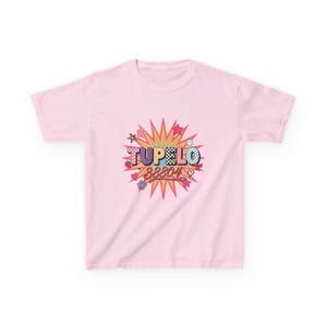 May include: A light pink short-sleeve t-shirt featuring a central graphic of a multi-coloured starburst. The text TUPELO 38804 is printed in the centre with various patterns including checkers, stripes, and polka dots, surrounded by small icons.