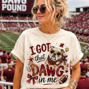 May include: A cream-colored short-sleeve t-shirt featuring a graphic print of a bulldog wearing heart-shaped sunglasses and a bandana. The text reads I GOT that DAWG in me, accompanied by illustrations of a football, a bow, stars, and paw prints.
