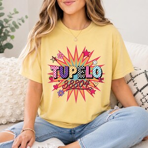 Op de afbeelding: Een lichtgeel t-shirt met korte mouwen en een kleurrijk grafisch ontwerp met de tekst TUPELO 38804. Het ontwerp bevat een grote sterrenvorm, ruitpatronen, stippen, strepen, kleine sterretjes, bloemen, een aardbei en een smiley.