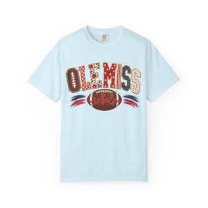 May include: A light blue short-sleeve t-shirt featuring the text OLE MISS in a multi-patterned red, white, and blue design. Below the text is a brown football graphic with the word rebels written in red script, flanked by red and blue brush strokes.