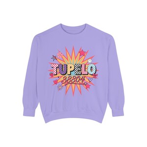 Op de afbeelding: Een lichtpaars sweatshirt met lange mouwen en een centrale afbeelding op een veelkleurige sterrenachtergrond. De tekst TUPELO 38804 staat in vetgedrukte letters, omringd door kleine iconen zoals sterren, hartjes, bloemen, een aardbei en een smiley.
