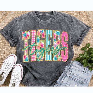 May include: A charcoal grey acid-wash t-shirt featuring the word TIGERS in large block letters with varied patterns including floral, pink gingham, teal stripes, and pink polka dots, with the word Tigers in teal cursive script overlaid.