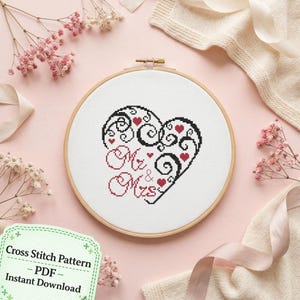 May include: A circular wooden embroidery hoop containing white fabric with a cross-stitch design of a black swirling heart, small red hearts, and the text "Mr & Mrs" in red. A green label with the text "Cross Stitch Pattern PDF Instant Download" is visible.