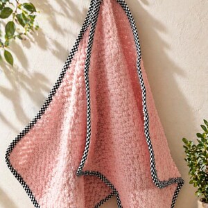 May include: A pink, fluffy towel with a black and white checkered border hangs from a hook on a light-colored wall. The towel's texture is soft and plush, and the checkered trim adds a classic touch. The towel is likely for use in a bathroom or spa setting.