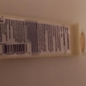 K&ouml;nnte beinhalten: Eine kleine, hellgelbe Tube mit wei&szlig;em Verschluss. Die Tube hat ein Etikett mit Text und einem Barcode. Das Produkt ist wahrscheinlich ein Kosmetik- oder K&ouml;rperpflegeartikel. Die Tube liegt auf einer hellen Oberfl&auml;che.