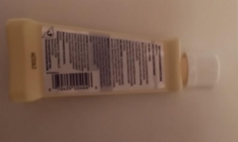 K&ouml;nnte beinhalten: Eine kleine, hellgelbe Tube mit wei&szlig;em Verschluss. Die Tube hat ein Etikett mit Text und einem Barcode. Das Produkt ist wahrscheinlich ein Kosmetik- oder K&ouml;rperpflegeartikel. Die Tube liegt auf einer hellen Oberfl&auml;che.